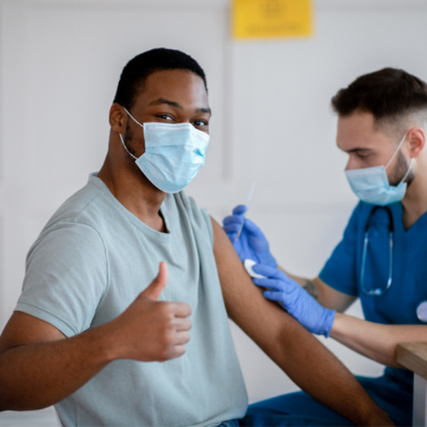 African American man in antiviral mask gesturing thumb up during coronavirus vaccination, approving of covid-19 immunization. Young doctor giving vaccine injection to male patient | Doylestown Health